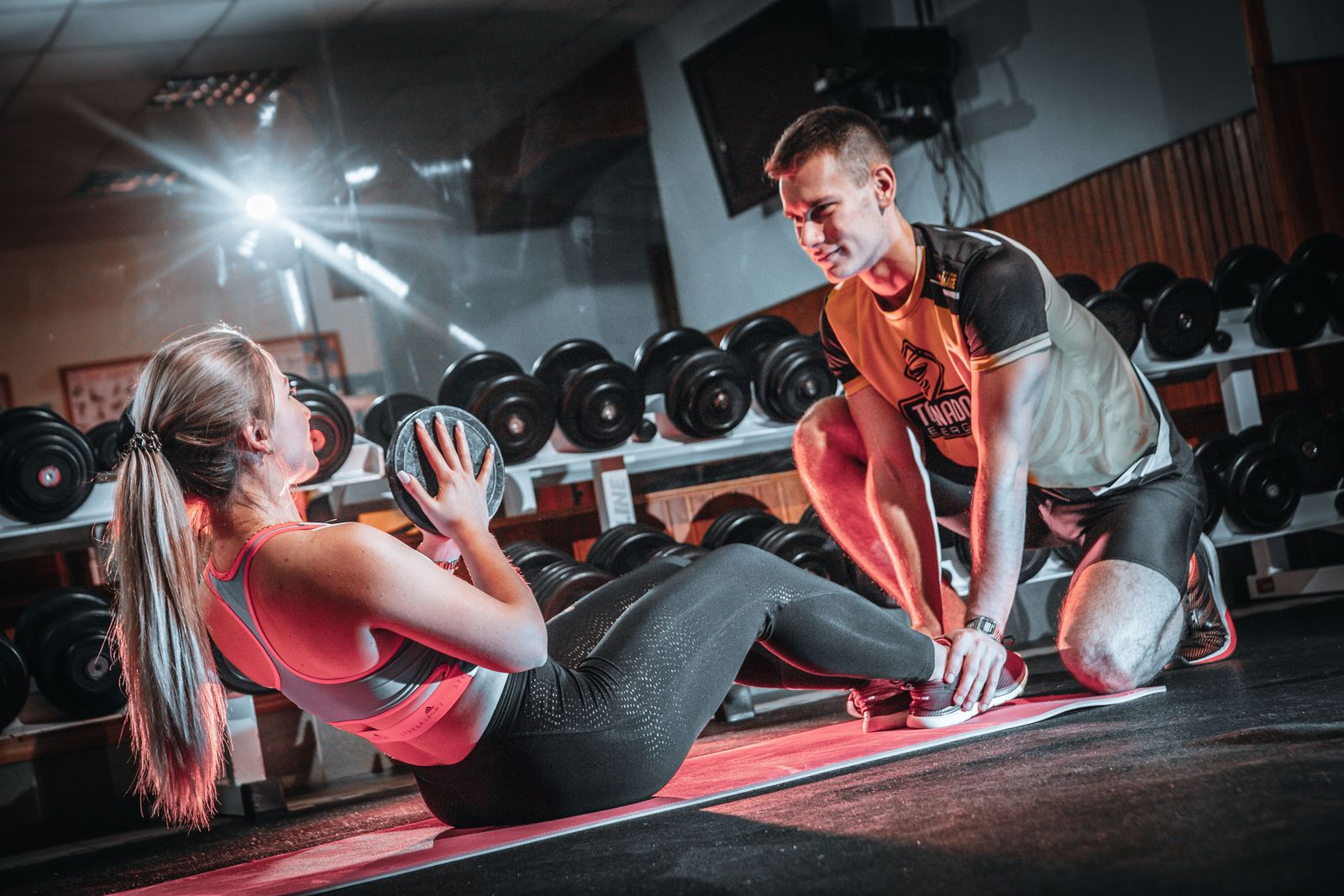 Woman performing a weighted sit-up holding a plate while a coach braces her feet on a mat